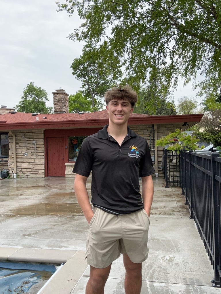 Young man in black polo shirt and khaki shorts standing in front of a mid-century home with red roof and stone exterior