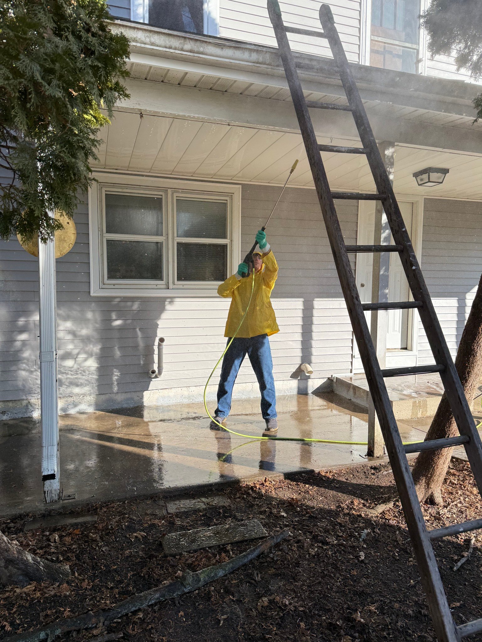 Person in yellow jacket power washing house patio with tall black ladder leaning against roof